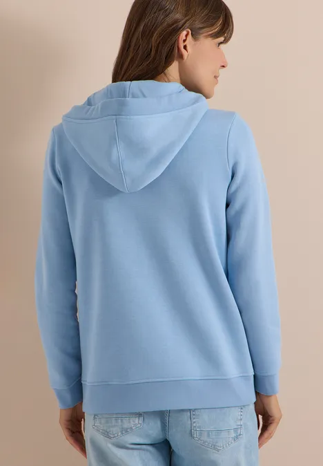 Frau von hinten im hellblauen Hoodie, lässig in Jeans vor neutralem Hintergrund.