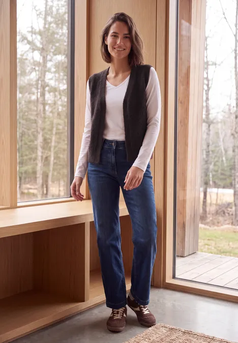 Eine Frau in Jeans, weißem Shirt und Weste steht lächelnd vor einem großen Fenster mit Blick auf Wälder.