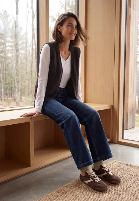 Eine junge Frau sitzt lächelnd auf einer Holzbank am Fenster, trägt Jeans, weißes Shirt und dunkelbraune Schuhe.