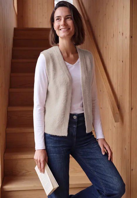 Eine lächelnde Frau in Jeans und Weste steht an einer Holztreppe und hält ein Buch.