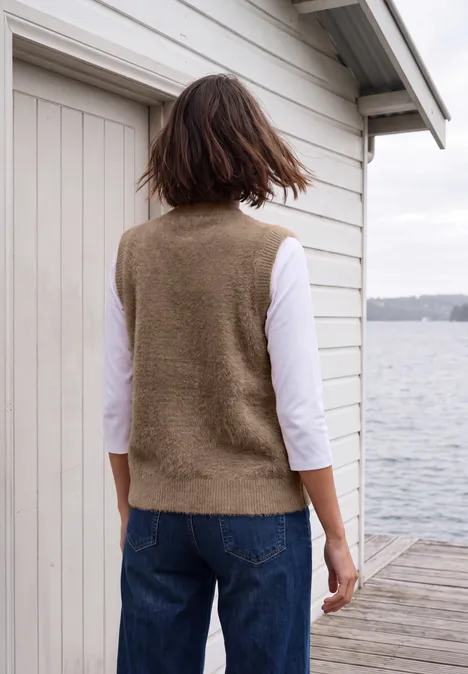 Eine Frau steht in Weste und Jeans auf einem Steg vor einem Bootshaus und blickt aufs Wasser.