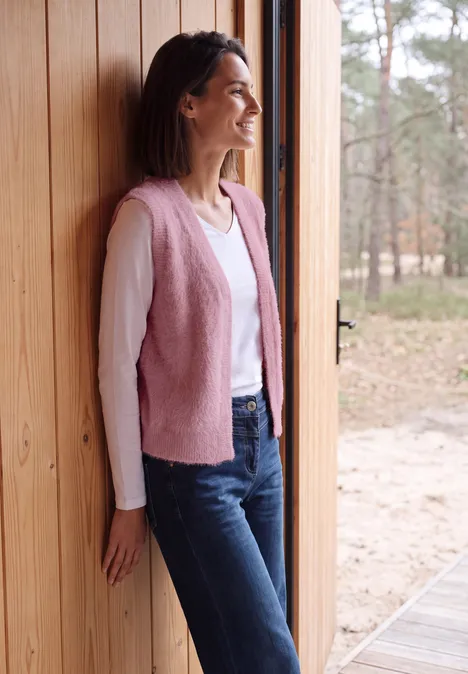 Eine Frau steht lächelnd in einer Holztüröffnung, trägt eine rosa Weste, weißen Pullover und Jeans. Wald im Hintergrund.