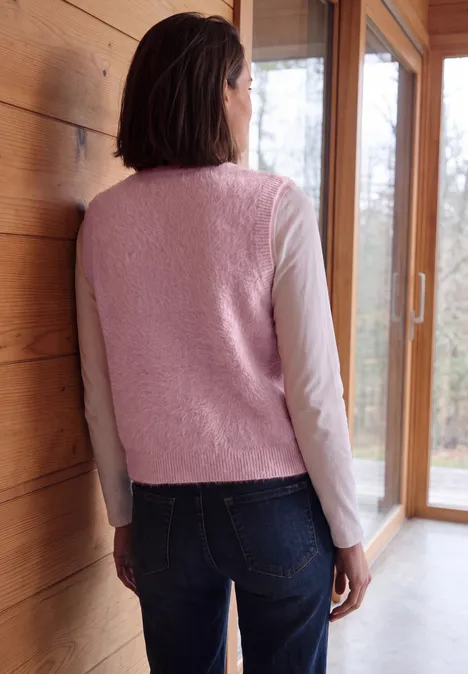 Frau steht mit dem Rücken zur Kamera, trägt rosafarbenen Fleece-Weste, Langarmshirt und Jeans in Holzhaus.