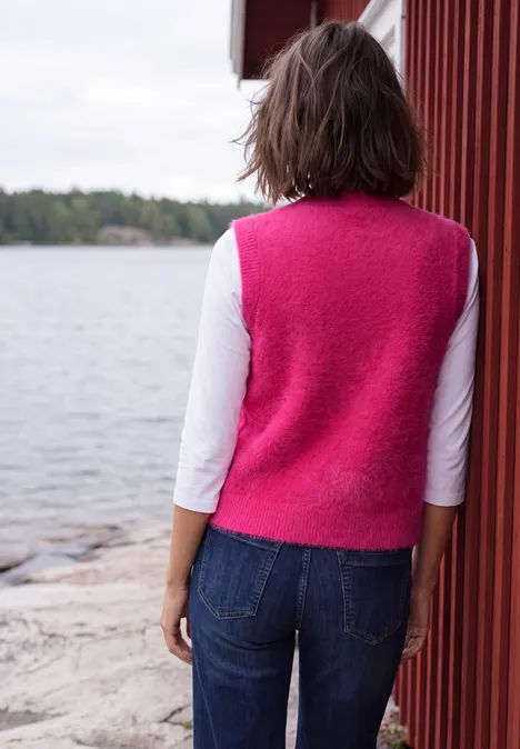 Eine Person steht mit dem Rücken zur Kamera in pinker Weste, weißem Shirt und Jeans an einem See.