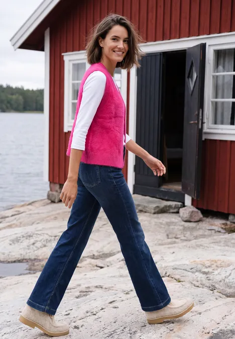 Frau in Jeans, weißem Shirt und pinker Weste läuft lächelnd am Ufer vor roter Hütte.
