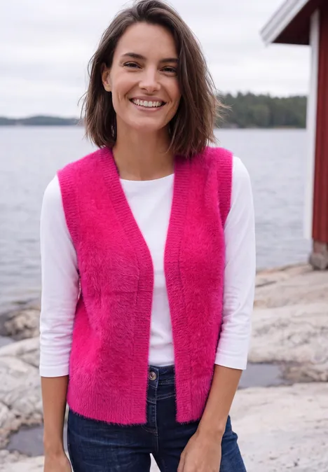 Eine lächelnde Frau in pinker Weste und weißem Shirt steht vor einem See mit rotem Bootshaus.