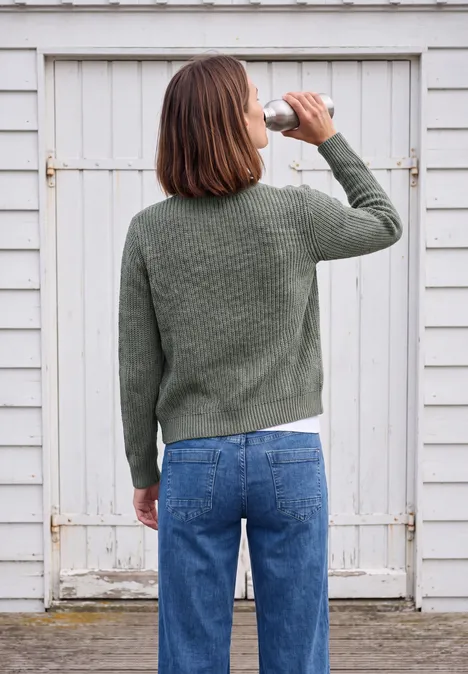Dikke vest met V-hals en gemêleerd effect easy khaki melange Eine Frau in grünem Pullover und blauer Jeans trinkt aus einer Flasche und steht vor einer weißen Holztür.