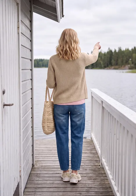 Dikke vest met V-hals en gemêleerde look creamy mocha melange Frau steht auf Holzterrasse mit Korbtasche, trägt Jeans und Pullover, schaut auf ruhigen See.