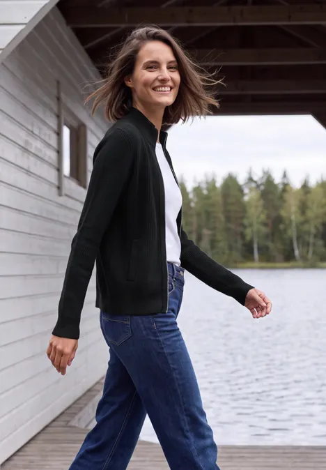 Vrouw in zwarte cardigan en jeans wandelt lachend langs een meer bij een houten chalet.