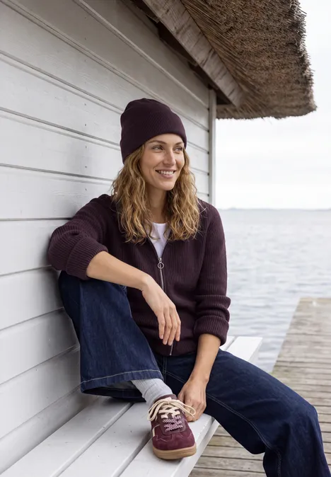 Vest met opstaande kraag en ritssluiting eggplant red melange Frau in lila Mütze und Pullover lehnt entspannt an einer weißen Strandhütte am Wasser und lächelt.