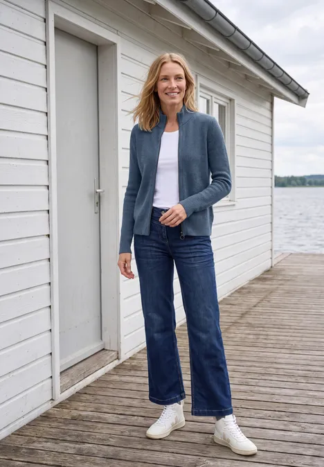 Vrouw in jeans, wit shirt en vest glimlacht op een steiger voor een wit boothuis aan het meer.