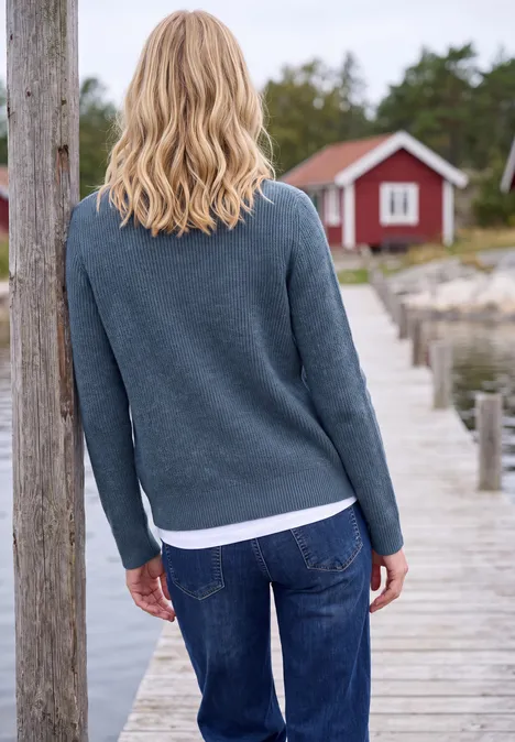 Een blonde vrouw staat op een houten steiger en kijkt naar een rood huisje aan het water.