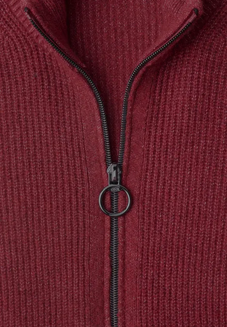 Close-up van een rood geribd gebreid kledingstuk met centrale rits en ronde ritsbedel.