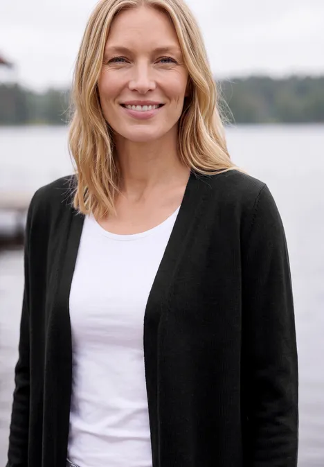 Lang vest met losse pasvorm Black Lächelnde Frau mit hellem Haar, weißem Shirt und dunkler Strickjacke vor ruhiger See-Kulisse.