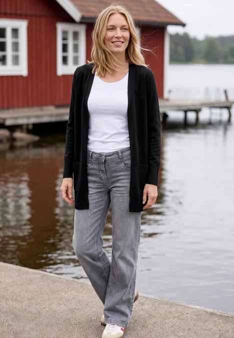 Lang vest met losse pasvorm Black Eine Frau in Jeans und Cardigan steht auf einem Steg vor einem roten Holzhaus am See.