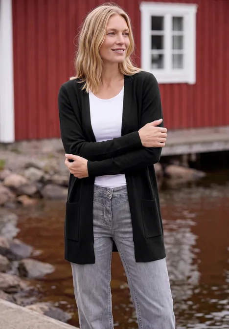 Lang vest met losse pasvorm Black Eine lächelnde Frau in schwarzer Strickjacke, weißem Top und grauen Jeans steht vor einem roten Holzhaus am Wasser.