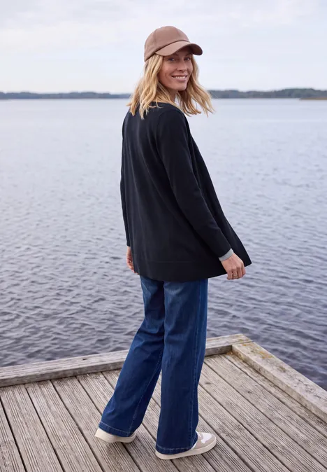 Długi kardigan o luźnym kroju urban dark blue Młoda kobieta w dżinsach, czarnej bluzie i czapce uśmiecha się na drewnianym pomoście nad jeziorem.