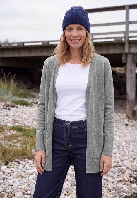 Długi kardigan o luźnym kroju sporty grey melange Uśmiechnięta kobieta w szarym kardiganie, białym T-shircie, dżinsach i niebieskiej czapce stoi swobodnie na kamienistej plaży przed drewnianym pomostem.
