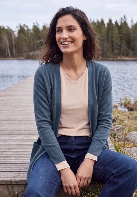 Lang vest met losse pasvorm cotton blue melange Frau sitzt lächelnd auf Holzsteg am See, trägt Cardigan und Jeans vor bewaldetem Hintergrund.