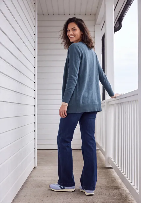 Lang vest met losse pasvorm cotton blue melange Frau in blauem Cardigan und Jeans schaut lächelnd über die Schulter auf weißer Terrasse.