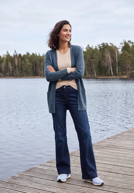 Lang vest met losse pasvorm cotton blue melange Frau steht entspannt auf einem Holzdock am See, trägt Jeans, Cardigan und Sneaker, Wald im Hintergrund.
