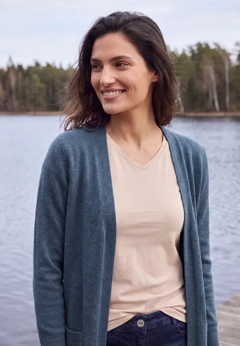 Lang vest met losse pasvorm cotton blue melange Eine lächelnde Frau am Seeufer trägt eine Strickjacke über einem hellen Shirt.