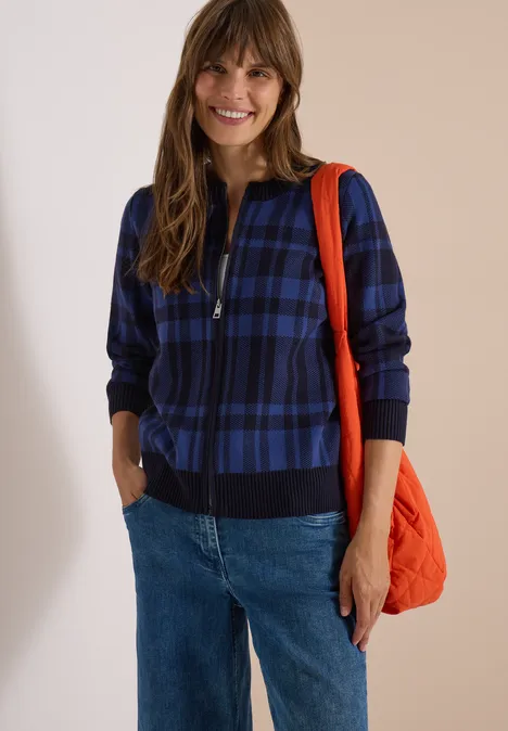 Frau lächelt, trägt eine blau-karierten Zip-Jacke, Jeans und eine orange Tasche.