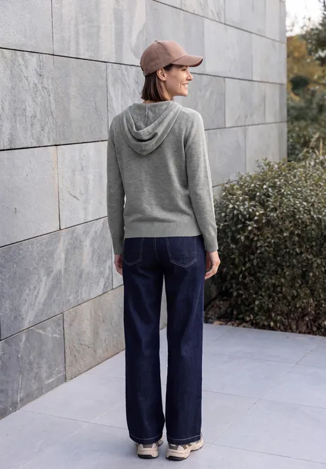 Cardigan à manches longues et à capuche flint grey melange Une personne en sweat à capuche gris, jean large et casquette se tient de profil devant un mur de pierre.