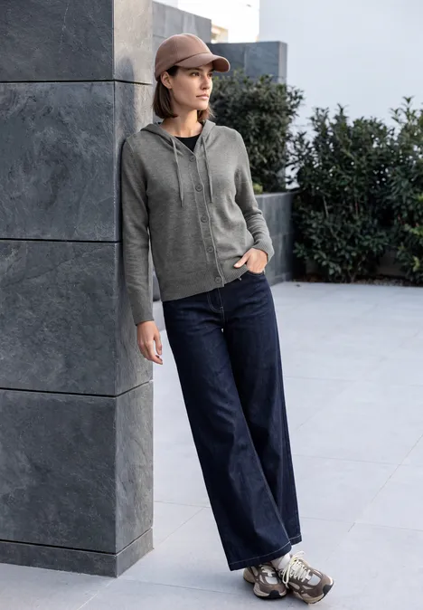 Cardigan à manches longues et à capuche flint grey melange Une femme en sweat à capuche gris, jean ample et casquette s’appuie nonchalamment contre un mur.