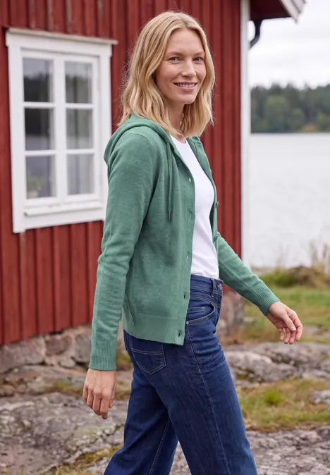 Een blonde vrouw in een groene capuchontrui en jeans lacht voor een rood houten huis aan een meer.