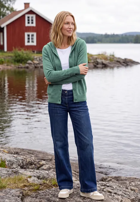 Vrouw staat op rotsen aan een meer, draagt groene cardigan, wit shirt en spijkerbroek, rood huis op achtergrond.
