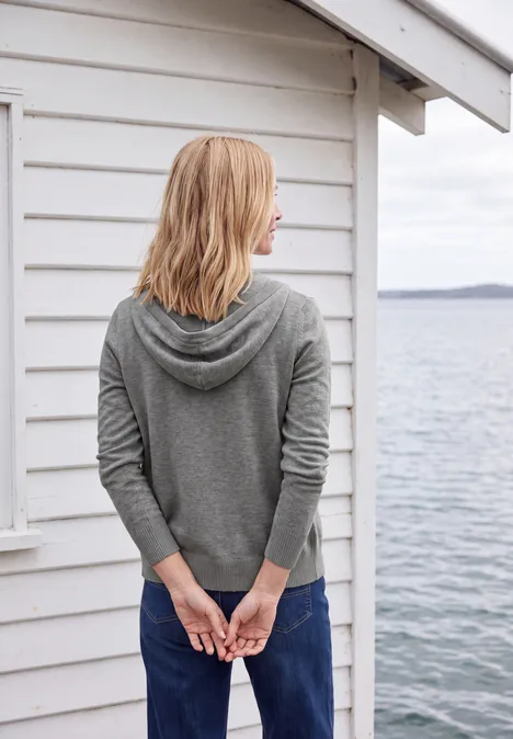 Vrouw in grijze hoodie, handen achter rug, voor een wit houten huisje aan het water.