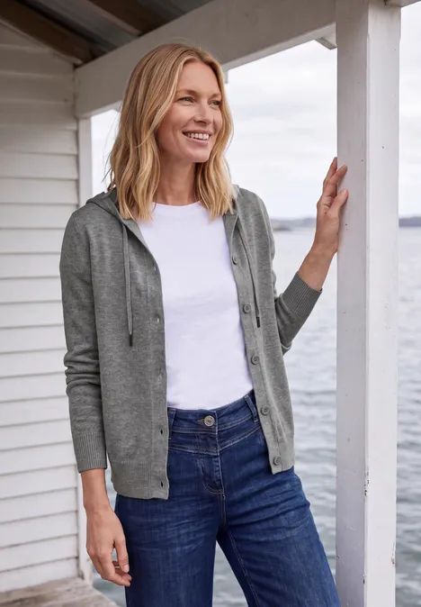 Glimlachende vrouw in grijze hoodie, wit T-shirt en blauwe jeans leunt tegen een wit houten huis aan zee