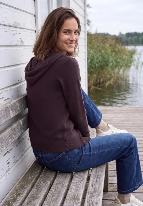 Langarm Cardigan mit Kapuze eggplant red melange Frau mit brauner Kapuzenjacke und Jeans sitzt seitlich auf einer Holzbank an einem See und lächelt.