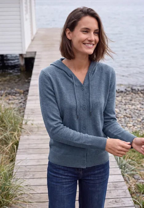 Eine Frau in Jeans und Kapuzenjacke steht auf einem Steg am Meer und lächelt.