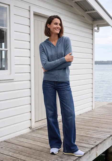 Eine lächelnde Frau in Jeans und Strickjacke steht auf einem Holzsteg vor einem weißen Haus am Wasser.