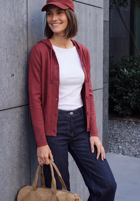 Cardigan à manches longues et à capuche rhubarb melange Femme souriante appuyée sur un mur, casquette et cardigan rouges, t-shirt blanc, pantalon foncé et sac beige.