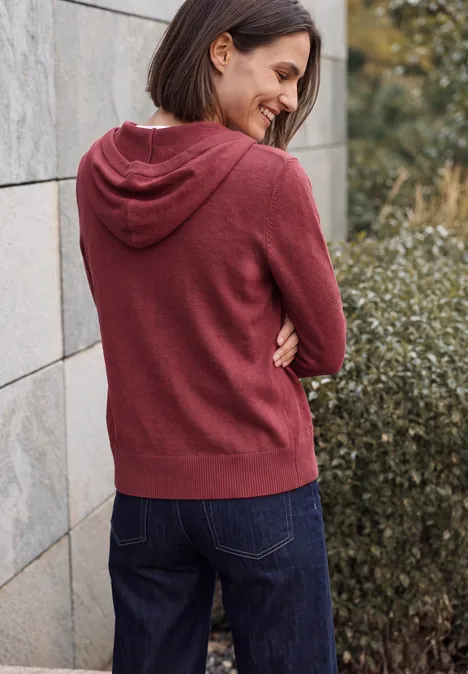 Cardigan à manches longues et à capuche rhubarb melange Femme souriante vue de dos, portant un sweat à capuche bordeaux et un jean, appuyée sur un mur près d’un buisson.