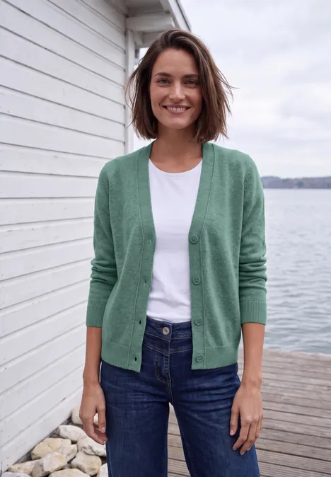 Frau in grüner Jacke, weißem Shirt und Jeans steht lächelnd auf einem Steg am See.