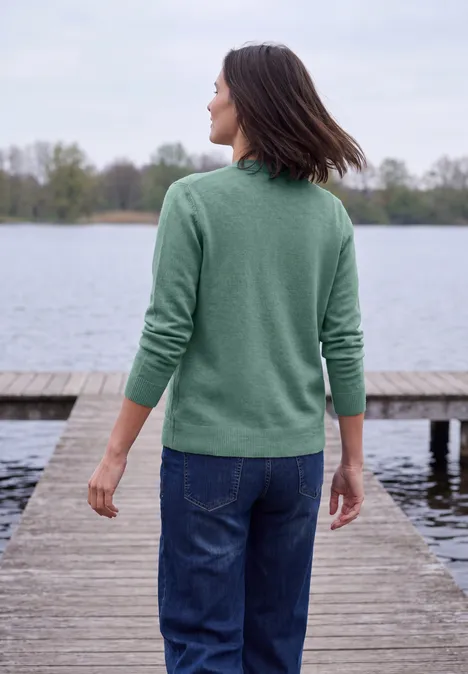 Frau in grünem Pullover und Jeans steht auf Steg und blickt auf ruhigen See.