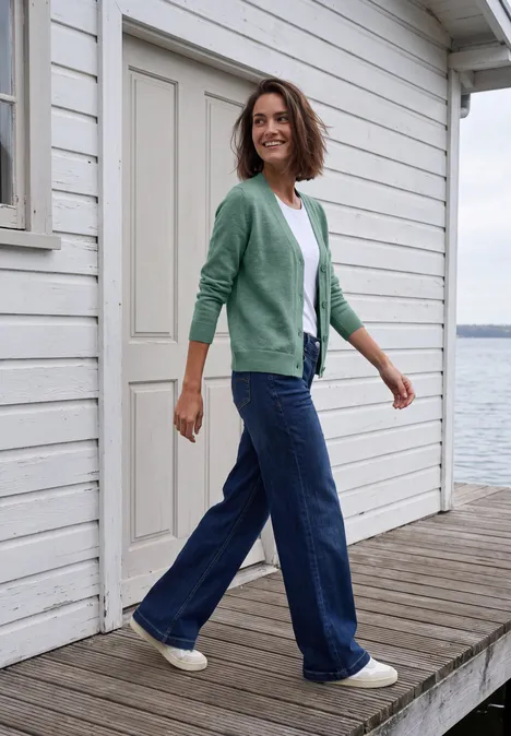 Frau in grünem Cardigan und Jeans spaziert auf einem Holzsteg vor weißem Haus am Wasser.
