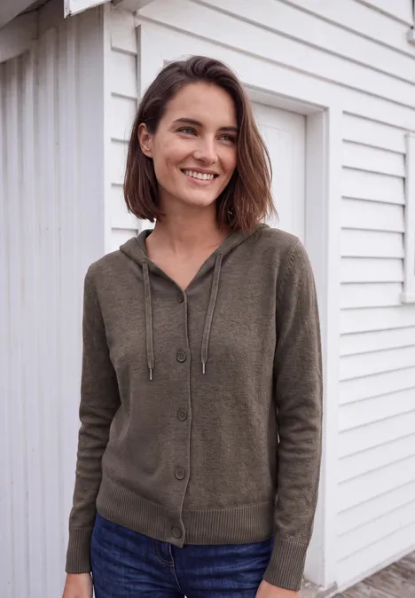 Eine lächelnde Frau in grünem Kapuzenpullover vor weißer Fassade.