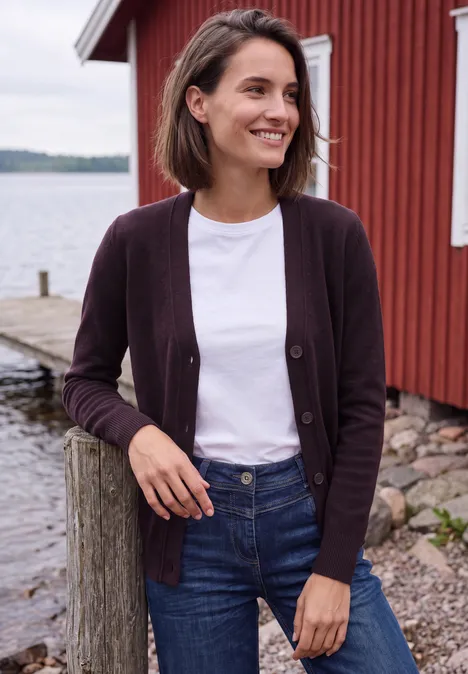 Frau in Jeans, weißem T-Shirt und violetter Strickjacke lächelt an einem Steg vor roter Holzscheune und Wasser.
