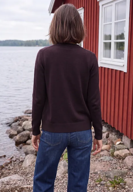 Eine Frau steht mit dem Rücken zur Kamera in Jeans und dunklem Pullover vor einem roten Haus am Wasser.