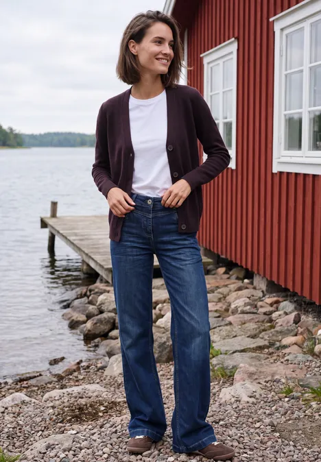 Frau steht in Jeans und Cardigan vor rotem Holzhaus am Seeufer.