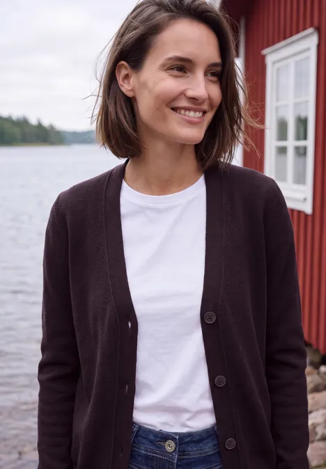 Eine Frau in schwarzer Strickjacke und weißem T-Shirt steht lächelnd vor einem roten Holzhaus am See.