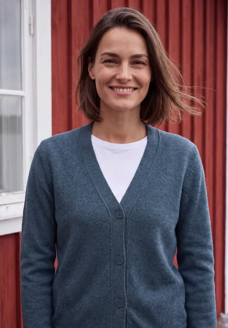 Une femme aux cheveux bruns courts sourit en cardigan gris devant une maison en bois rouge.