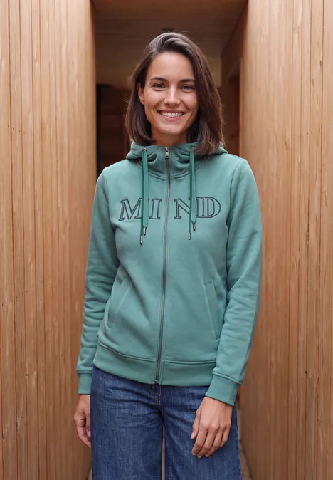 Sweatjacke mit Wording Artwork mineral blue Eine lächelnde Frau in grünem Kapuzenpullover mit 'MIND'-Schriftzug vor hölzerner Wand.