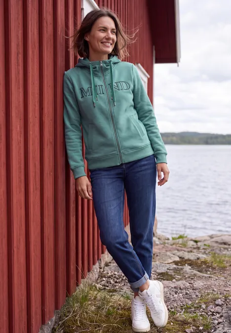 Sweatjacke mit Wording Artwork mineral blue Eine Frau lehnt an einer roten Holzwand am Seeufer, trägt eine türkisgrüne Kapuzenjacke und Jeans und lächelt.