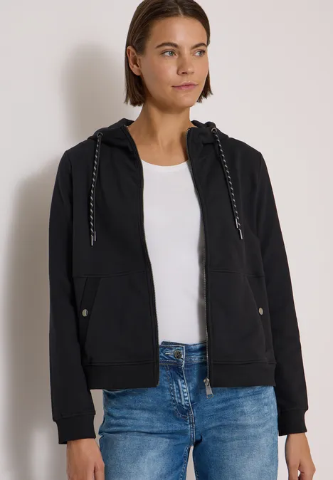 Frau trägt schwarze Kapuzenjacke, weißes T-Shirt und Jeans.